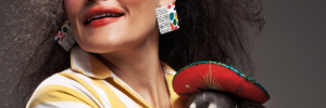 A profile picture of Jibz Cameron with rainbow eye makeup, color graphic earrings, holding a chihuahua wearing a tiny sombrero.